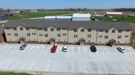 Parkside Manor in Haubstadt, Indiana: the three storey apartment building with beige siding above a brick base, gabled entrances along the front, its parking lot in the foreground and farm fields behind it.